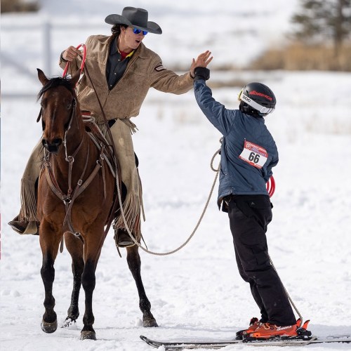 A cowboy on horseback interacts with a skier on the ground, both in a snowy outdoor setting.