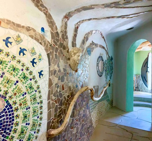 A colorful mosaic hallway with intricate designs and a sculpted face, leading to a doorway in a serene setting.