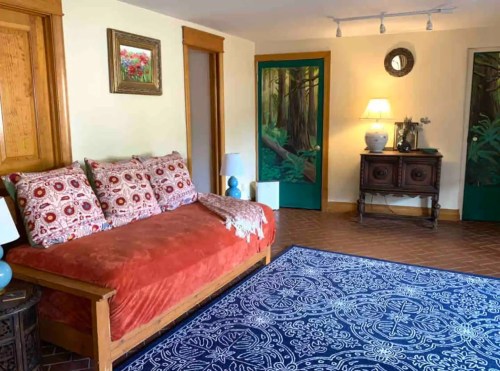 Cozy living room with a red daybed, decorative pillows, a blue rug, and forest-themed doors. Warm lighting adds ambiance.