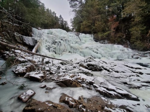 A frozen waterfall surrounded by snow-covered rocks and trees, creating a serene winter landscape.