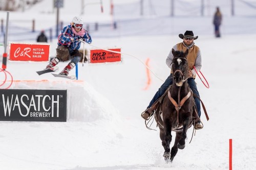 A cowboy on horseback chases a snowboarder jumping over a snow ramp at a winter event. Snowy landscape in the background.