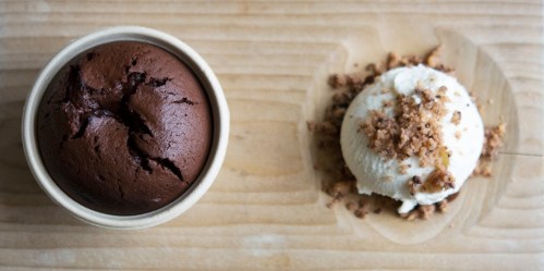 A rich chocolate lava cake in a bowl next to a scoop of vanilla ice cream topped with crumbled cookies.