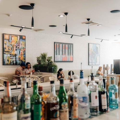 A modern bar scene with colorful artwork, patrons enjoying drinks, and a variety of bottles on the counter.