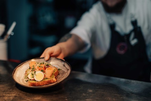 A chef presents a plate of toast topped with smoked salmon and garnished with herbs, set in a dimly lit kitchen.
