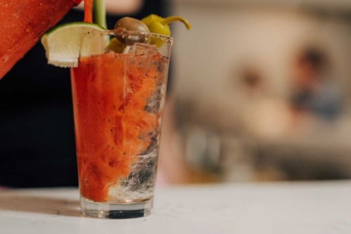 A close-up of a glass filled with a red cocktail, garnished with olives, lime, and a pepper.
