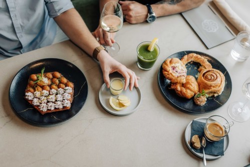 A table with three plates of food, including pastries, a green drink, and a glass of white wine.