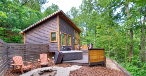 A cozy wooden cabin surrounded by trees, featuring a deck, hot tub, and outdoor seating area.