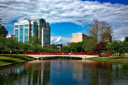 A serene park scene featuring a pond, a red bridge, and modern buildings under a blue sky with fluffy clouds.