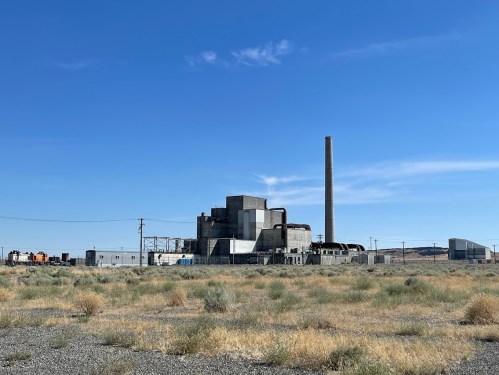 Industrial facility with a tall smokestack, surrounded by dry grass and clear blue sky.