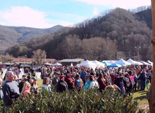 A large crowd gathers at an outdoor event with tents, surrounded by mountains and trees under a clear blue sky.