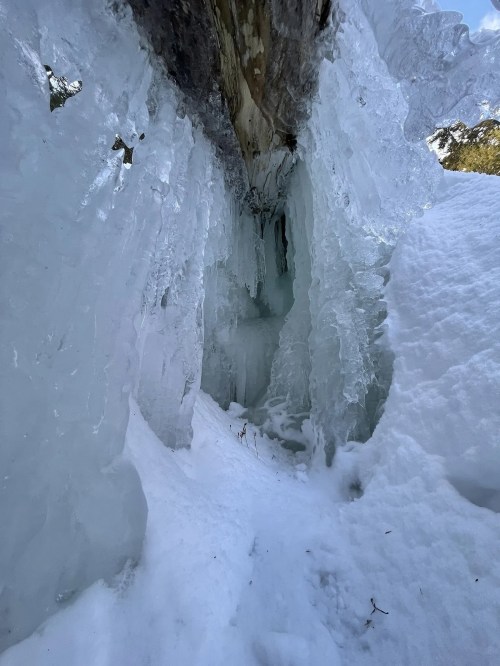 Icicles hang from a rocky overhang, surrounded by snow, creating a wintery cave-like scene.
