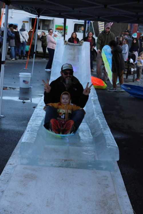 A man and a child slide down an ice slide at a festive outdoor event, surrounded by onlookers and colorful equipment.