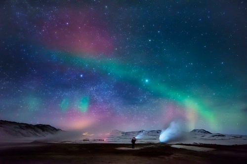 A person stands under a vibrant night sky filled with stars and colorful auroras over a snowy landscape.