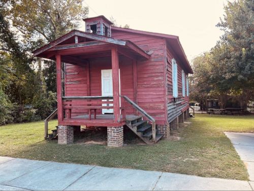 A red wooden house with a porch and steps, surrounded by grass and trees, on a sunny day.