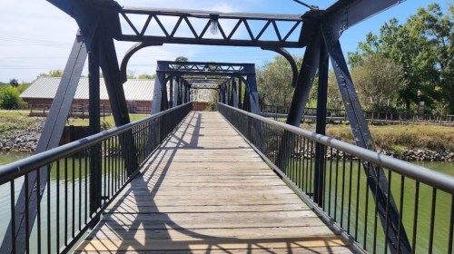 A wooden walkway on a metal bridge over a green river, surrounded by trees and buildings in the background.