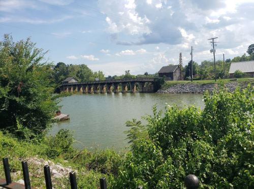 A serene river scene with a bridge, greenery, and a small boat near an old building under a partly cloudy sky.