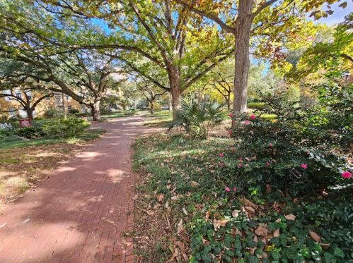 A brick pathway winds through a lush park with trees, shrubs, and colorful flowers under a clear blue sky.