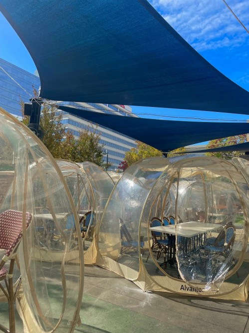 Clear outdoor dining pods with tables, shaded by a blue canopy, against a backdrop of buildings and a bright sky.
