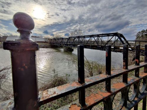 A rusty fence in the foreground frames a bridge over a calm waterway under a cloudy sky.