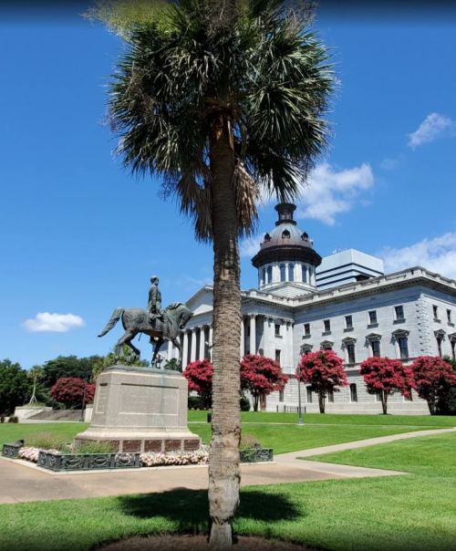 A statue of a horseman stands in front of a historic building, surrounded by palm trees and blooming flowers under a blue sky.