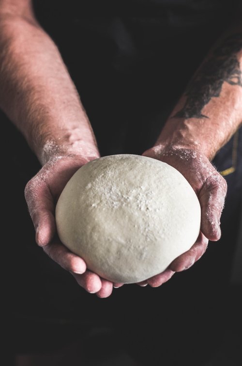 A person holds a round ball of dough, hands dusted with flour, against a dark background.
