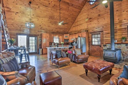 Cozy cabin interior with wooden walls, leather furniture, a kitchen area, and a stone fireplace. Bright and inviting atmosphere.