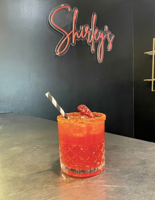 A vibrant red cocktail with a striped straw and chili garnish, set against a black wall with "Shirley's" sign.