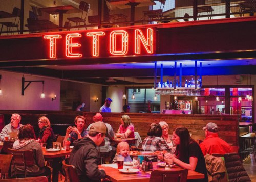 A lively restaurant scene with diners at wooden tables, a neon "TETON" sign, and a bar in the background.