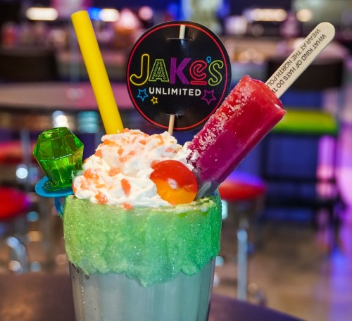 A colorful milkshake topped with whipped cream, candy, and a sign for Jake's Unlimited, set against a vibrant background.