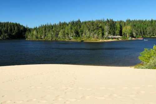 A sandy beach leads to a calm lake surrounded by lush green trees and a distant shoreline with cabins.