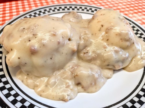 A plate of two fluffy biscuits smothered in creamy sausage gravy, served on a checkered tablecloth.