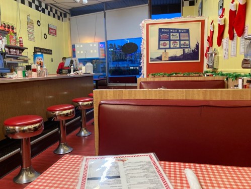 A retro diner interior with red booths, chrome stools, and festive decorations, featuring a menu on a checkered tablecloth.