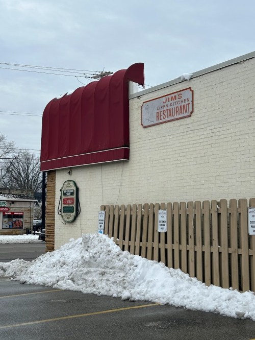 Exterior of Jim's Open Kitchen Restaurant, featuring a red awning and a snow pile in front, on a cloudy day.