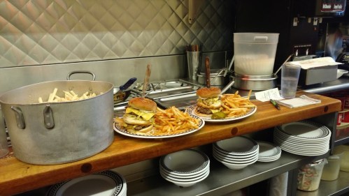 A kitchen counter with two plates of burgers and fries, a pot of fries, and stacked plates in the background.