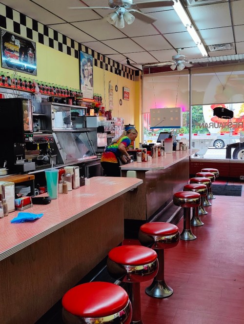 A retro diner interior with red stools, a counter, and a staff member in a tie-dye shirt preparing food.