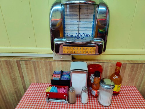 A vintage jukebox on a wall above a checkered tablecloth with condiments, napkins, and utensils arranged neatly.