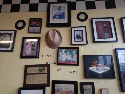 A wall display featuring framed photos, a hat, and memorabilia labeled "Jim's Hall of Fame" in a diner setting.