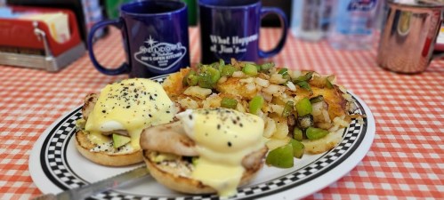 A plate of eggs Benedict with hash browns, topped with green peppers, served with two coffee mugs in the background.