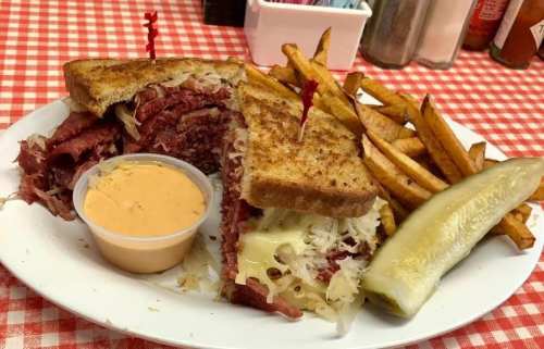 A plate featuring a corned beef sandwich with sauerkraut, fries, a pickle, and a side of dipping sauce.