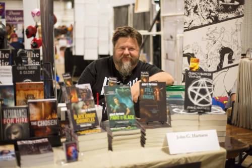 A bearded man sits behind a table filled with books at a convention, smiling at the camera.