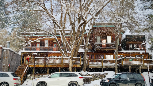 A snowy scene of a wooden cabin with multiple balconies, surrounded by trees and parked cars in winter.