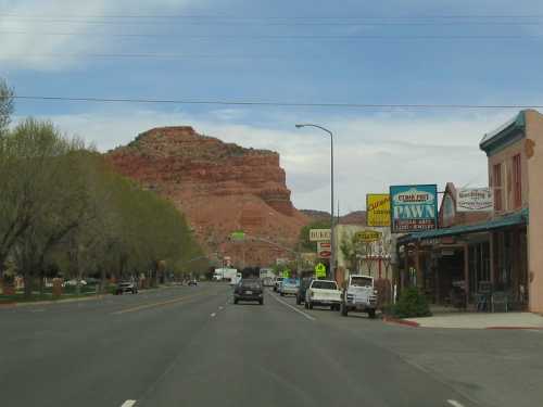 A scenic view of a road lined with shops, leading to a large red rock formation under a blue sky.