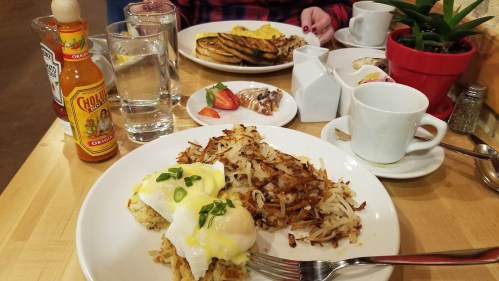 A breakfast table featuring eggs Benedict, hash browns, coffee, and a bottle of hot sauce, with a plant in the background.
