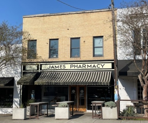 A brick building with a sign reading "James Pharmacy" and outdoor seating under a striped awning.