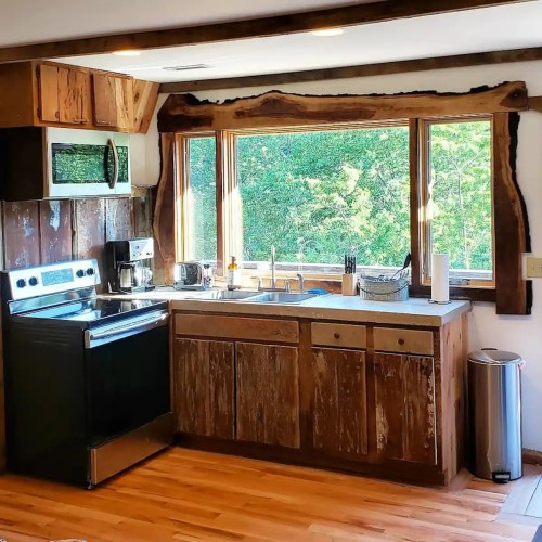 A rustic kitchen with wooden cabinets, stainless steel appliances, and large windows overlooking greenery.