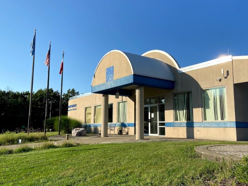 A modern building with a curved roof, surrounded by flags and greenery, under a clear blue sky.
