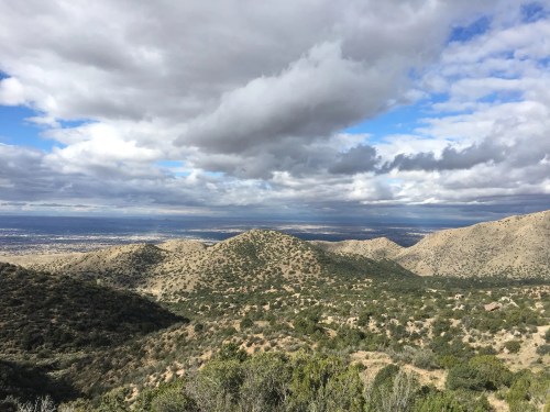 A panoramic view of rolling hills and valleys under a cloudy sky, with distant mountains and a hint of a city below.