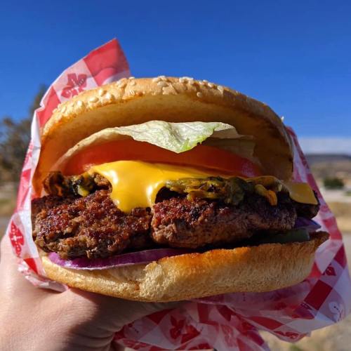 A hand holding a juicy cheeseburger with lettuce, tomato, and pickles against a clear blue sky.