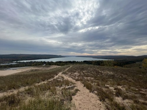 A scenic view of a lake surrounded by hills and trees under a cloudy sky, with a sandy path in the foreground.