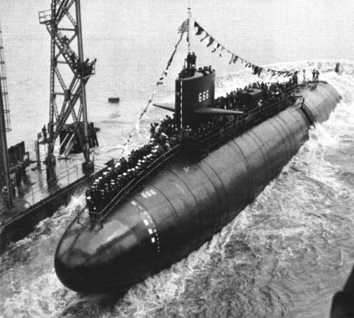 A black and white photo of a submarine, with crew members on deck, launching from a dock adorned with flags.